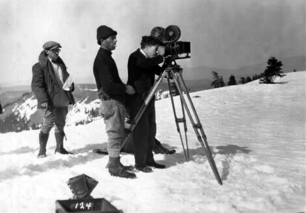 Charlie Chaplin operando uma câmera em The Gold Rush enquanto outros membros da equipe ficam perto dele — Foto: Wikimedia Commons